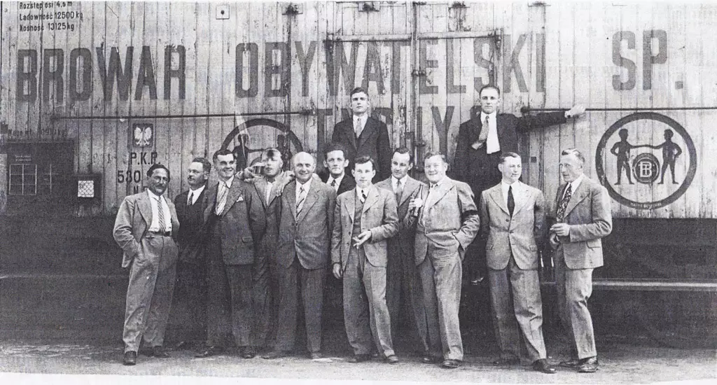 Grupa mężczyzn w garniturach pozująca przed budynkiem browaru Obywatelskiego.